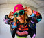 Person in colorful jacket and Disco Tribe shirt poses playfully, wearing a pink beanie with a yellow star and teal sunglasses, in a bright industrial space.
