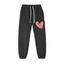 Black jogger pants with a pink broken heart graphic on the left thigh. The heart has a yellow lightning bolt and the letters D and T inside. White drawstring at the waistband.