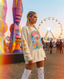 Woman with braids and glitter makeup wears a graphic sweatshirt and white lace-up boots at a colorful outdoor festival with art sculptures and a Ferris wheel in the background.