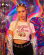 A young woman with blonde braided hair wears The Bender Crop—a white festival tee featuring the phrase "GET IN LOSER WE'RE GOING ON A BENDER" and a camper graphic. She completes her look with black jeans, a celestial belt, and a metallic jacket, capturing the perfect music festival vibe in a neon, disco-themed studio.