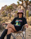 A woman wearing a pink bucket hat, sunglasses, the Disco With the Tribe Tee, and boots sits on a folding chair in a desert landscape with rocks and Joshua trees in the background.