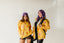 Two women wearing yellow and purple patterned jackets and black shorts pose together against a white background.