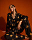 A person poses against an orange background, wearing the Midnight Stardust Silk Set (Bottoms)—black satin pants adorned with large yellow stars—paired with hoop earrings and braided hair, one arm resting on their knee.