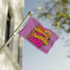 A small purple “I am Literally Just a Festival Girl Flag” from the Electric Forest Totem and Festival Campsite Decor collection features colorful, playful lettering with whimsical doodles of mushrooms, a rainbow, and stars. The flag is displayed outdoors on a pole near a tree and a white building.