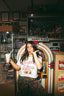 A woman poses playfully in front of a jukebox, holding a milkshake. She wears The Bender Crop with patterned pants. The setting is a retro diner filled with nostalgic decorations and vintage memorabilia.