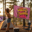 At sunset among tall pine trees, a woman stands near her tent at a campsite, holding the “I am Literally Just a Festival Girl Flag,” a vibrant pink Electric Forest Totem and festival campsite decor featuring colorful lettering.