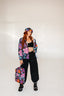 A woman stands in a studio wearing black pants, boots, a black crop top, and a matching floral jacket, complemented by the Electric Petals Bucket Hat and a backpack with colorful flower patterns.