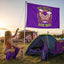 A woman in colorful festival attire kneels beside a tent, holding the “Festival Campsite Flag | Festival Totem | Bad Bitches Love Bass Flag,” which features a butterfly and floral design. A music stage and crowd appear in the background, capturing the vibrant spirit of a bass music festival.