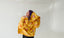 A person with purple hair wearing a yellow hooded jacket with orange sunburst patterns is smiling and looking up.
