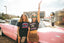 Two women stand beside a pink vintage car. One, smiling with her arm raised and sunglasses on, flaunts the Hot Girls Love IndieTronica Crop Top. Both display festival fashion in black graphic tees and skirts. A modern building and a retro diner complete the sunny backdrop.