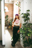 A woman wearing The Bender Crop, paired with a purple headscarf, black pants, and chunky shoes stands indoors by a concrete pillar adorned with green plants in a room featuring brick walls and large windows.