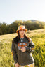 A woman stands in a sunny, grassy field wearing the Electric Tiger Crewneck—an oversized sweatshirt featuring a bold tiger graphic—paired with a floral bucket hat and patterned pants, making for an effortlessly cool festival look. Trees and a hill are visible in the background.
