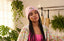A woman with long braids wears the DT Bolt Purp Edition Beanie and a floral jacket, standing indoors near green plants and a clothing rack.