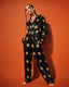 The person stands barefoot against an orange background, wearing the Midnight Stardust Silk Set bottoms—black satin pants adorned with yellow stars. Their hands rest in their pockets as they showcase long braids, large hoop earrings, and bold eye makeup.