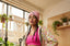 A person with long braids wears the DT Bolt Purp Edition Beanie, paired with a vibrant floral jacket and a pink crop top, standing in a sunlit room adorned with hanging plants and macrame decor near large windows.
