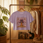 A purple crop top with a mushroom graphic and the text "Disco Tribes" is hanging on a clothing rack in a room with various clothing items and plants.