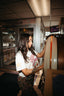 Wearing The Bender Crop, a woman with long dark hair and a tattoo on her left arm stands beside a jukebox in a retro-style diner, choosing a song as sunlight streams through the windows and casts a warm glow inside.