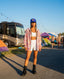 A woman in shorts, sports bra, patterned jacket, and boots stands at an outdoor festival with tents and an RV in the background. She wears the StarGaze Trucker – Purple Haze hat with an adjustable snapback closure and looks directly at the camera, perfectly embodying effortless festival fashion.