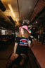 A smiling woman with curly hair, wearing a pink hat and the Hot Girls Love IndieTronica Crop Top, sits at a diner counter holding a milkshake and looking to the side.