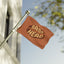 A Bass Head Festival Flag New, featuring the words BASS HEAD in bold yellow letters on a brown background, waves on a pole outside a building, resembling a classic rave flag or festival totem, with tree branches and a blue sky in the background.