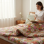 A woman is sitting on a floral-patterned bed, reading a book and sipping a beverage.