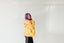 A woman with purple hair is standing in front of a white background, wearing a yellow jacket with a floral pattern.