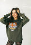 A woman, smiling with her hands in her long dark hair, models an oversized olive-green Electric Tiger Crewneck adorned with a bold tiger’s face, paired with patterned black lace leggings—her festival look styled flawlessly against a plain white background.