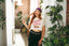 A woman with red hair stands indoors near plants, wearing The Bender Crop—a festival crop tee that reads "Get in loser, we are going on a bender"—paired with black pants featuring a chain belt and a purple headscarf, creating a perfect music festival outfit.
