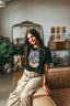 A woman with long brown hair leans on a couch, wearing a black Epic Playlist Society Crop Top and patterned pants. A mirror, plants, and records are visible in the cozy, stylish room background.