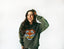 Wearing the Electric Tiger Crewneck—an oversized green sweatshirt with a bold graphic—a woman with long hair stands against a white background, smiling with her eyes closed and one hand touching her head, capturing effortless festival clothing inspiration.