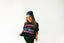 Someone wearing a colorful bandana holds out the Hot Girls Love IndieTronica Crop Top—a black crop top with bold, vibrant lettering—against a plain white background, making it an ideal choice for fans of music-inspired clothing and festival fashion.
