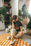 A smiling woman with long hair sits on a patterned rug by a mirror, wearing the Epic Playlist Society Crop Top and patterned pants for the perfect festival outfit. Chunky sneakers complete her look, while disco balls and bookshelves fill the background, accented by nearby plants.