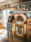 Wearing the Epic Playlist Society Crop Top, a woman in a blue cap and checkered pants stands next to a vintage jukebox in a retro diner filled with nostalgic memorabilia and posters, her outfit seamlessly blending classic festival vibes with timeless retro flair.