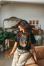 Wearing the Epic Playlist Society Crop Top and patterned pants, a woman with long brown hair twirls joyfully on a couch—her festival-ready look shining in a stylish, cozy room filled with plants, a mirror, and eclectic decor.