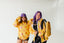 Two women wearing yellow and purple patterned tops and black pants pose together against a white background.
