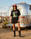 Wearing the Disco With the Tribe Tee, a woman stands in front of an Airstream trailer in a desert setting, sporting sunglasses and a pink bucket hat. String lights and colorful flags set the backdrop, perfectly capturing laid-back festival fashion.