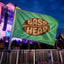 A Bass Music Festival Flag waves boldly at a music festival, its striking design capturing the spirit of the night. In front of a vibrant stage lit with colorful lights, a lively crowd gathers—some raising their EDM Festival Totem or Bass Head Totem for Raves & Camping high above as they celebrate together amidst the energy of basshead camp decor.