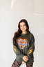 A woman with long dark hair wears the Electric Tiger Crewneck, an oversized sweatshirt showcasing a vibrant tiger graphic and striking yellow lightning bolts. She pairs it with lace-patterned leggings for a bold festival-ready look, standing against a white background and smiling with her hands folded in front.