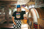 A woman wearing a blue cap, sunglasses, the Epic Playlist Society Crop Top, and checkered pants stands in a retro diner beside a jukebox, surrounded by neon signs and vintage decor.