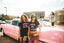 Two women pose in front of a vintage pink car, embracing festival style. One wears a black skirt and graphic tee, while the other sports green sunglasses and the Hot Girls Love IndieTronica Crop Top—an eye-catching piece that channels music-inspired clothing vibes. A building and diner can be seen in the background.