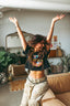 A woman with long hair raises both arms joyfully in a cozy, stylish living room. She wears the Epic Playlist Society Crop Top, perfectly channeling festival outfit vibes, paired with patterned pants and smiling with her eyes closed.