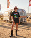 Wearing the "Disco With the Tribe Tee," black boots, a pink hat, and sunglasses, a woman stands on sandy ground in front of a silver Airstream trailer adorned with colorful flags, effortlessly embodying festival style.