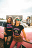 Two women showcase their festival fashion in front of a pink vintage car. One wears green sunglasses, a black Leaving the Afters shirt, leopard skirt, and pink fishnets—bringing music-inspired clothing to life. The other rocks the Hot Girls Love IndieTronica Crop Top with curly hair for the ultimate cool-girl vibe.