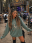 A woman with long, braided hair smiles and poses at an outdoor music festival, wearing the cozy Celestial Harmony Crewneck featuring a sun, moon, and star design. Behind her, the festival stage and crowd are visible among tall trees.