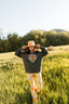 Standing in a grassy field with trees and a hill behind them, a person wears the oversized Electric Tiger Crewneck featuring its distinctive green tiger-print design, paired with colorful floral pants and a matching cap—making a bold statement in festival attire.