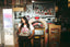 A woman stands by a jukebox, sipping a drink with a straw. She wears The Bender Crop and patterned pants in a retro-style diner filled with vintage decor and collectibles.