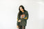 Wearing the Electric Tiger Crewneck—an olive green oversized sweatshirt with a striking tiger graphic and yellow lightning bolts—a person with long dark hair pairs it with black lace leggings for eye-catching festival style against a plain white background.