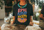 A person wearing the Epic Playlist Society Crop Top—a black graphic tee with colorful "EPIC PLAYLIST SOCIETY" text—paired with a bold necklace, sitting indoors surrounded by disco balls and plants. The perfect inspiration for your next festival outfit!.