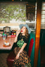 Wearing the Disco Trip Tee, a woman with a blue hat and leopard print pants sits smiling in a retro diner booth, enjoying a cherry. On the table beside her are an ice cream dessert, napkins, and a jukebox.