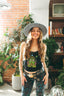 A smiling woman in a wide-brimmed hat and the Astral Connection Festie Tank stands indoors, surrounded by paisley pants, plants, books, and a disco ball.